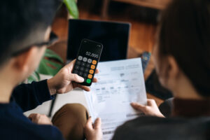 Young Asian Couple Managing Personal Banking And Finance At Home. Planning Budget And Calculating Expenses While Checking Bills With Calculator. Managing Taxes, Financial Bills And Online Stock Trades With Laptop. Home Finances And Budgeting Concept