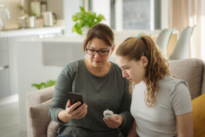 Portrait Of Mother And Teenager Daughter Using Phone And Glocometer To Check Blood Sugar Level Of The Girl
